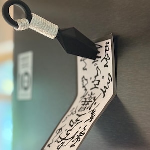 Könnte beinhalten: Ein schwarzer Magnet in Kunai-Form mit einem weiß umwickelten Griff, der einen rechteckigen weißen Zettel mit abstrakten schwarzen Tintenmarkierungen an einer metallischen Oberfläche befestigt. Der Magnet hat einen Ring am Griffende.