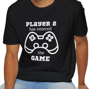 May include: Black t-shirt with white text and a controller graphic. The text reads "PLAYER 2 has entered the GAME". The design features a stylized video game controller. The shirt is made of a soft material.