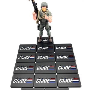 May include: A G.I. Joe action figure holding a machine gun, standing above a collection of black rectangular bases. Each base features the G.I. Joe logo with red, white, and blue stripes. The figure is dressed in a military uniform.
