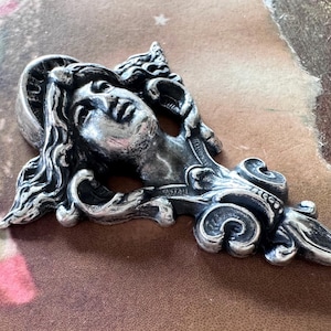 May include: A silver-toned pendant featuring a classical female face with flowing hair and ornate detailing. The pendant has a halo and is set against a vintage-style background with floral and brown tones. The design is intricate and detailed.