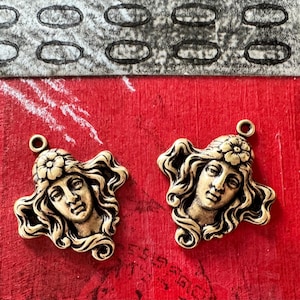 May include: Two antique gold-colored metal pendants. Each pendant features a woman's face with flowing hair and a flower crown. The pendants are on a red surface with a patterned background.