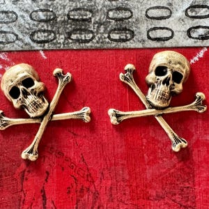 May include: Two gold-toned skull and crossbones charms. The skulls have black eye sockets and are detailed with a textured surface. The crossbones are made of thin metal and are crossed at the center.