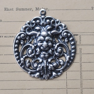 May include: Silver pendant with an ornate design featuring a grotesque face in the centre. The pendant is circular and has a loop at the top for hanging.