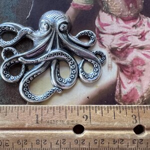 K&ouml;nnte beinhalten: Ein silberfarbener Anh&auml;nger in Form eines Oktopus mit detaillierten Tentakeln und einem strukturierten Kopf. Der Anh&auml;nger wird neben einem Holzlineal pr&auml;sentiert. Der Hintergrund zeigt ein Bild im Vintage-Stil mit Rosa- und Braunt&ouml;nen.