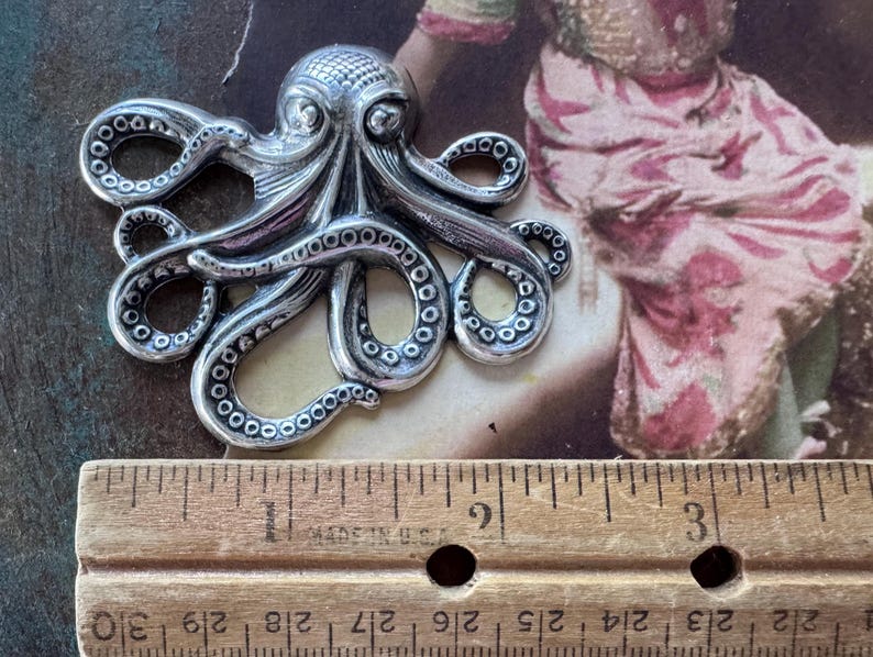 K&ouml;nnte beinhalten: Ein silberfarbener Anh&auml;nger in Form eines Oktopus mit detaillierten Tentakeln und einem strukturierten Kopf. Der Anh&auml;nger wird neben einem Holzlineal pr&auml;sentiert. Der Hintergrund zeigt ein Bild im Vintage-Stil mit Rosa- und Braunt&ouml;nen.