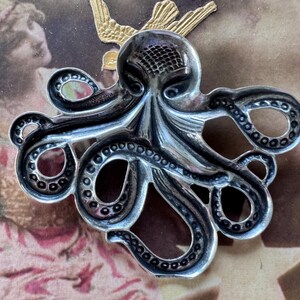 K&ouml;nnte beinhalten: Silberfarbene Oktopus-Brosche mit detaillierten Tentakeln und einem strukturierten Kopf. Die Brosche ist vor einem Hintergrund im Vintage-Stil mit einem goldfarbenen Vogelakzent platziert.