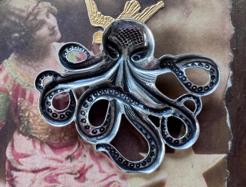 K&ouml;nnte beinhalten: Silberfarbene Oktopus-Brosche mit detaillierten Tentakeln und einem strukturierten Kopf. Die Brosche ist vor einem Hintergrund im Vintage-Stil mit einem goldfarbenen Vogelakzent platziert.