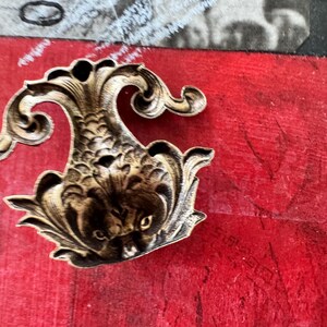 Puede incluir: Un adorno decorativo de metal dorado en forma de pez estilizado con detalles intrincados. El adorno tiene una superficie texturizada en relieve y una cola rizada.