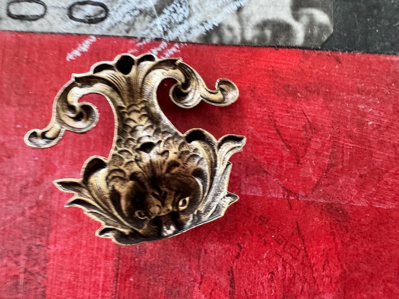 Puede incluir: Un adorno decorativo de metal dorado en forma de pez estilizado con detalles intrincados. El adorno tiene una superficie texturizada en relieve y una cola rizada.