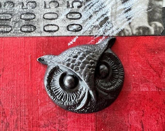 May include: A black metal owl figurine with detailed features, including a textured surface and large, round eyes. The owl is mounted on a red background.