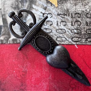 May include: A black metal sword with a heart-shaped blade and a decorative handle. The sword is on a red background.