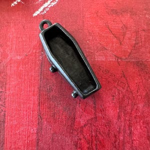 May include: A small, black metal coffin pendant with a hinged lid. The pendant is detailed with a textured surface and has a loop at the top for attaching a chain.
