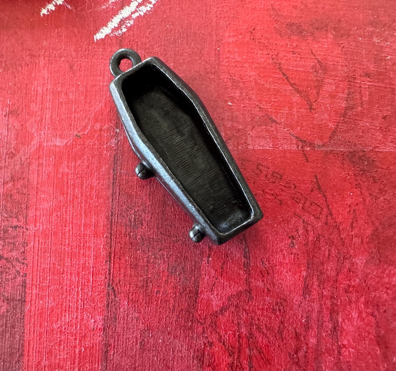 May include: A small, black metal coffin pendant with a hinged lid. The pendant is detailed with a textured surface and has a loop at the top for attaching a chain.