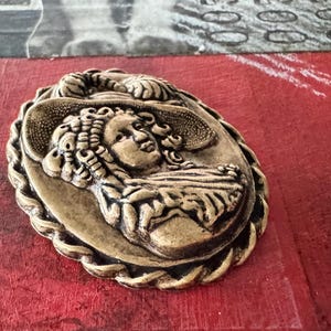 May include: A gold-toned metal cameo brooch featuring a woman with a large hat and elaborate hair. The cameo is set in a rope-like border.