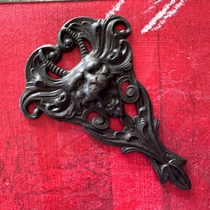 May include: A black metal decorative piece with a detailed design featuring a stylized face and intricate floral patterns. The piece is shaped like a heart and has a pointed bottom.