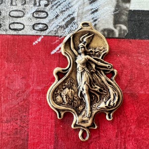 May include: A gold-toned metal pendant featuring a detailed relief sculpture of a woman in a flowing robe, holding a staff. The pendant has a unique, asymmetrical shape and a loop at the top for hanging.