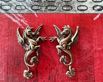 Op de afbeelding: Twee goudkleurige metalen bedels met gevleugelde paarden met gedetailleerde kenmerken. De paarden staan in een opgerichte houding met uitgespreide vleugels. De bedels hebben een decoratief, versierd ontwerp.