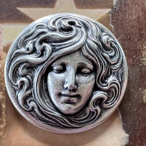 May include: A round, silver-toned metal button with a detailed relief of a woman's face. The woman has flowing hair and closed eyes. The button has a vintage or antique aesthetic.