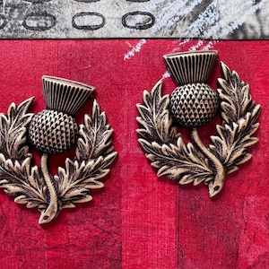 May include: Two gold-toned metal thistle charms with detailed leaf and flower designs. The thistle is a traditional Scottish symbol.