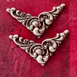 May include: Two ornate, antique-style metal corner embellishments. Each piece features a symmetrical design with floral and scrollwork details. The embellishments are a muted gold color, set against a textured, deep red background.