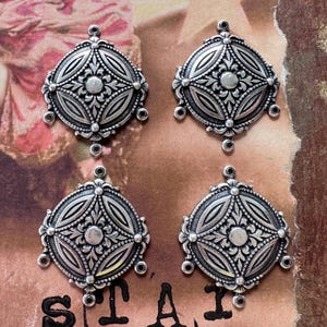May include: Four silver-coloured, ornate pendants with a circular design. Each pendant features a central floral motif surrounded by detailed patterns and small, dangling loops. The pendants are arranged in a square formation.