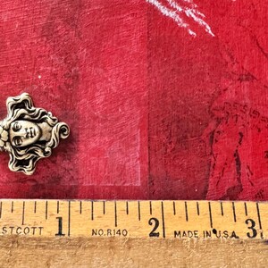 May include: A detailed, antique-style metal charm in the shape of a woman's face with flowing hair and floral accents. The charm is approximately 1 inch in length. The background is a textured red surface.