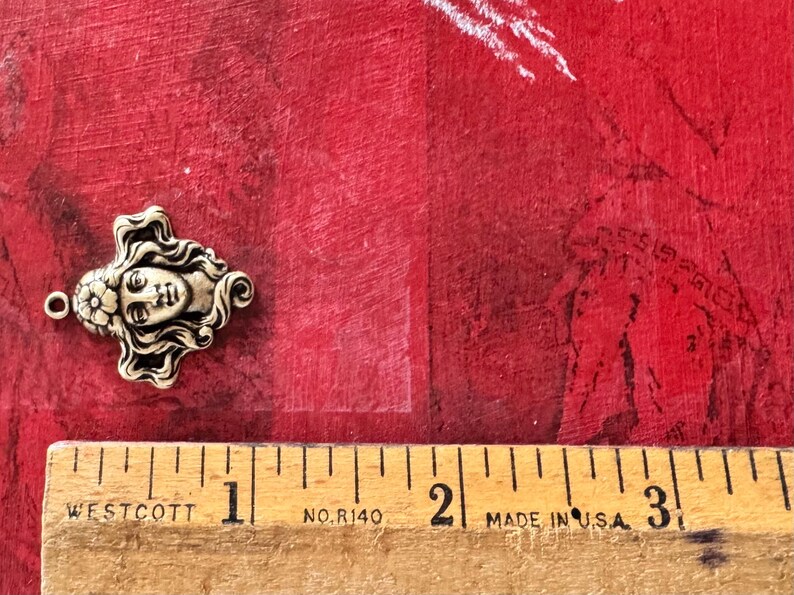 May include: A detailed, antique-style metal charm in the shape of a woman's face with flowing hair and floral accents. The charm is approximately 1 inch in length. The background is a textured red surface.