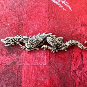 Op de afbeelding: Een zilverkleurig drakenbeeldje met gedetailleerde schubben en klauwen. De draak bevindt zich in een rennende houding met zijn kop naar de zijkant gedraaid.