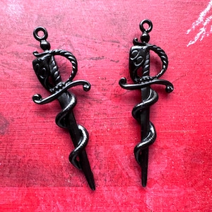 May include: Two identical matte black metal sword pendants, each featuring a coiled snake wrapping around the blade and a detailed hilt with a circular loop at the top for hanging. The sword designs are symmetrical with a pointed tip.