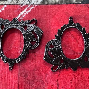 May include: Two ornate, black metal oval frames with intricate floral and scrollwork details. The frames are set against a textured red background, showcasing their decorative design. These frames are ideal for crafting or adding a vintage touch to artwork.