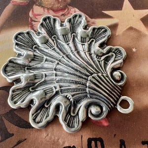May include: A detailed, silver-toned metal pendant with an ornate, shell-like design. The pendant features a series of curved, textured elements radiating from a central point, with a small loop for attachment. The background is a vintage-style image.