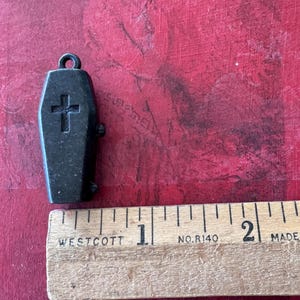 May include: A black metal coffin charm with a cross on the front. The charm is approximately 2.5 cm long and is hanging from a silver chain.