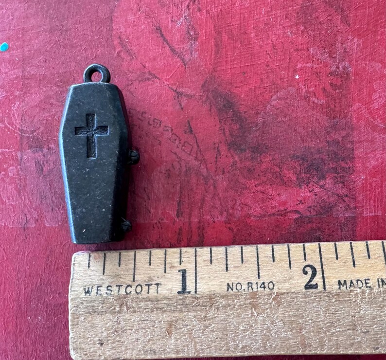 May include: A black metal coffin charm with a cross on the front. The charm is approximately 2.5 cm long and is hanging from a silver chain.