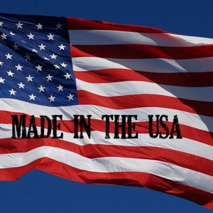 May include: A red, white and blue American flag waving in the wind against a blue sky. The flag has 13 red and white stripes and 50 white stars on a blue field. The text "MADE IN THE USA" is printed in black on the flag.