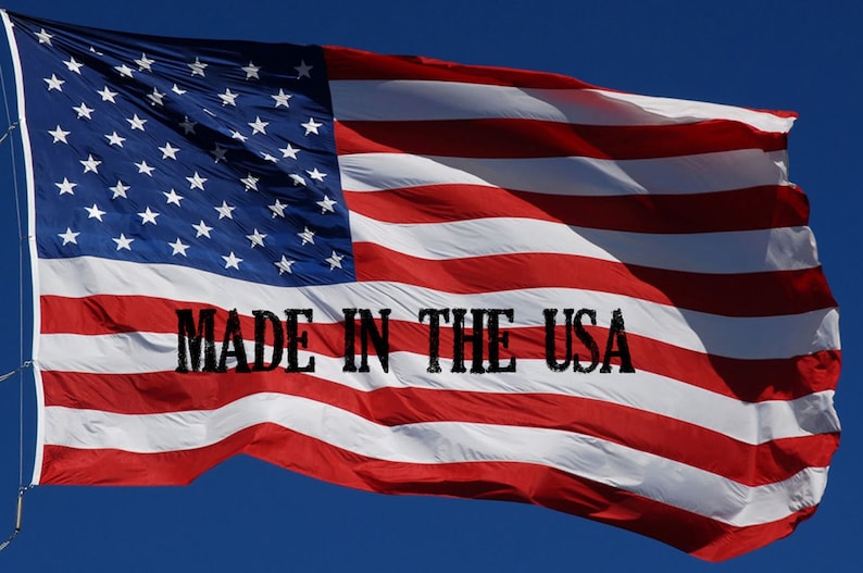 May include: A red, white and blue American flag waving in the wind against a blue sky. The flag has 13 red and white stripes and 50 white stars on a blue field. The text "MADE IN THE USA" is printed in black on the flag.