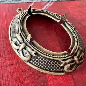 May include: A gold-toned metal oval frame with a decorative design of bows and floral motifs. The frame is open on the inside, suggesting it is for a picture or other decorative item.