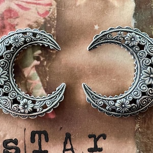 May include: Two silver crescent moon charms with intricate floral designs. The charms are made of metal and have a detailed filigree pattern.