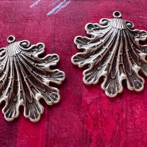 May include: Two ornate, antique-style metal pendants in a leaf or shell design. Each pendant is a detailed, openwork design with a loop at the top for hanging. The pendants are a bronze color and rest on a red surface.