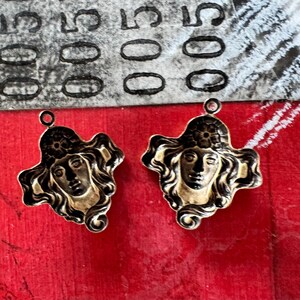 May include: Two gold-toned metal pendants featuring a woman's face with flowing hair and a floral headpiece. The pendants have a black outline and a loop at the top for hanging. The background is red.
