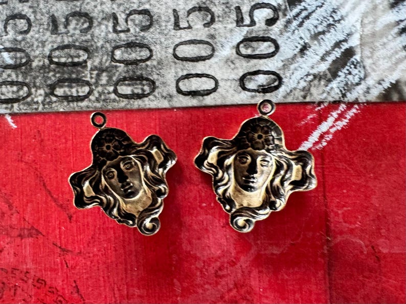 May include: Two gold-toned metal pendants featuring a woman's face with flowing hair and a floral headpiece. The pendants have a black outline and a loop at the top for hanging. The background is red.