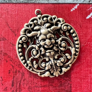 May include: A gold-toned metal pendant featuring a detailed design of a grotesque face surrounded by ornate scrollwork.