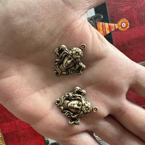 May include: Two antique bronze-colored metal charms. Each charm features a detailed profile of a woman's face with flowing hair and a floral accent. The charms have a small loop at the top for attaching to jewelry or other crafts.
