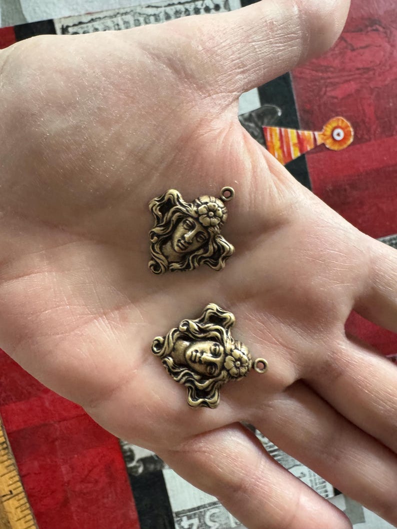 May include: Two antique bronze-colored metal charms. Each charm features a detailed profile of a woman's face with flowing hair and a floral accent. The charms have a small loop at the top for attaching to jewelry or other crafts.