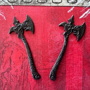 May include: Two black metal axe-shaped keychains with intricate floral designs. The keychains are on a red background.