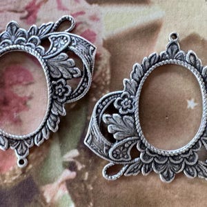 Könnte beinhalten: Zwei verzierte, silberfarbene ovale Anhängerrahmen mit floralen und Blattmotiven. Die Rahmen haben eine detaillierte, Vintage-Ästhetik mit einer offenen Mitte zum Einsetzen eines Fotos oder Designs. Jeder Rahmen hat eine Schlaufe zum Aufhängen.