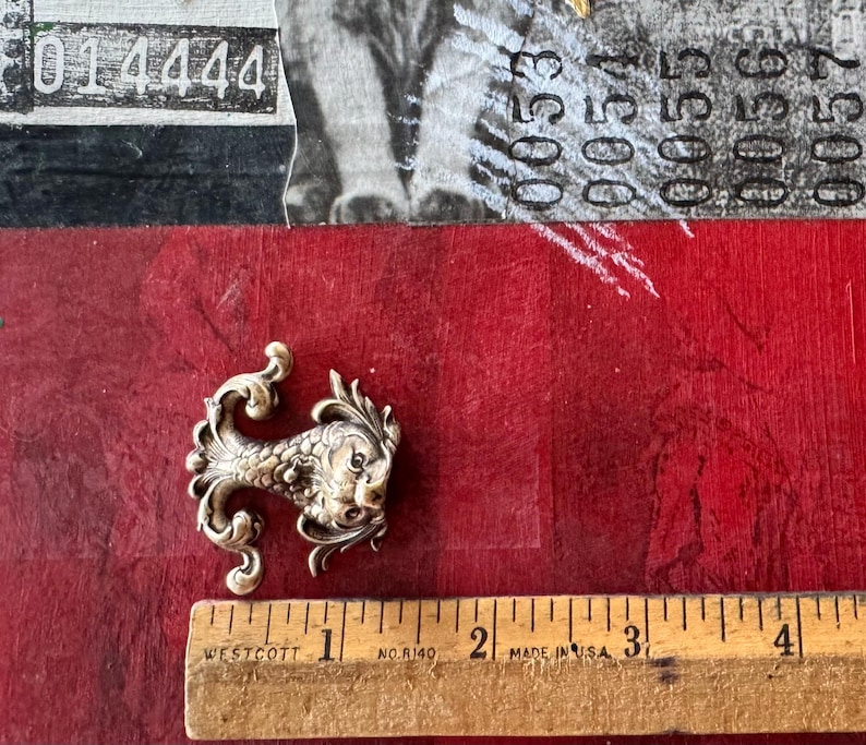 Puede incluir: Una figurita de pez de metal plateado con detalles intrincados. El pez tiene una cola rizada y una cara detallada. La figurita mide aproximadamente 5 cm de largo.