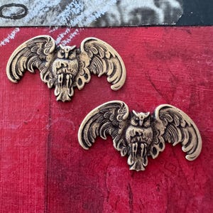 May include: Two antique gold-colored owl embellishments with outstretched wings. The detailed metalwork features a central owl figure with intricate feather and wing designs. The owls are set against a textured red background.