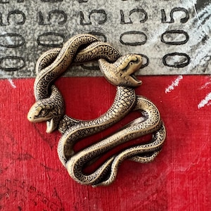 May include: A gold-toned metal snake charm in the shape of two snakes intertwined in a circle. The snakes have detailed scales and are facing each other.