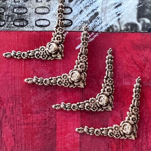 Peut inclure: Quatre ornements d'angle de couleur laiton antique avec des motifs floraux. Chaque pièce d'angle présente une rose centrale entourée de diverses fleurs et feuilles. Les pièces sont disposées sur une surface rouge texturée.
