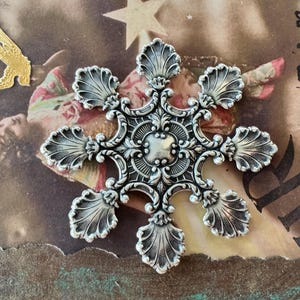 May include: A silver snowflake ornament with a detailed, ornate design. The snowflake has a scalloped edge and a raised center.